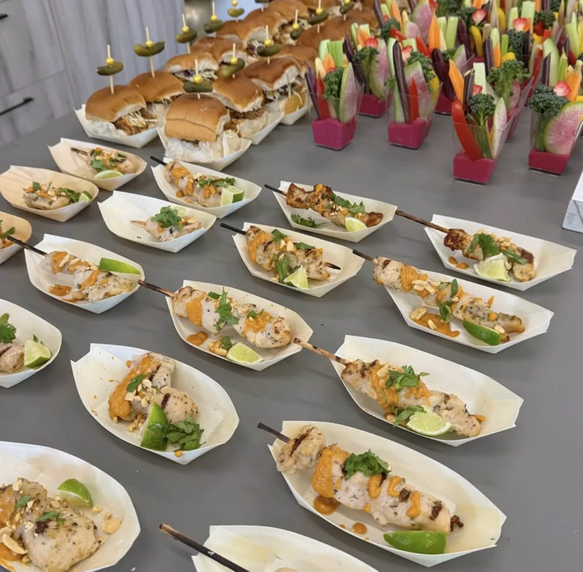 A table with various appetizers, including grilled chicken skewers with lime wedges, and small sandwiches with pickles. In the background, there are colorful vegetable arrangements and small sandwiches with decorative picks.