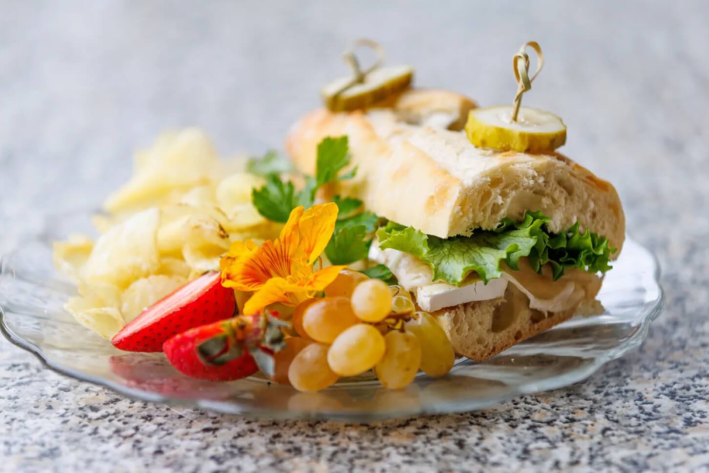 A plate with a club sandwich, grapes, strawberries, potato chips, and a flower garnish on a granite countertop.