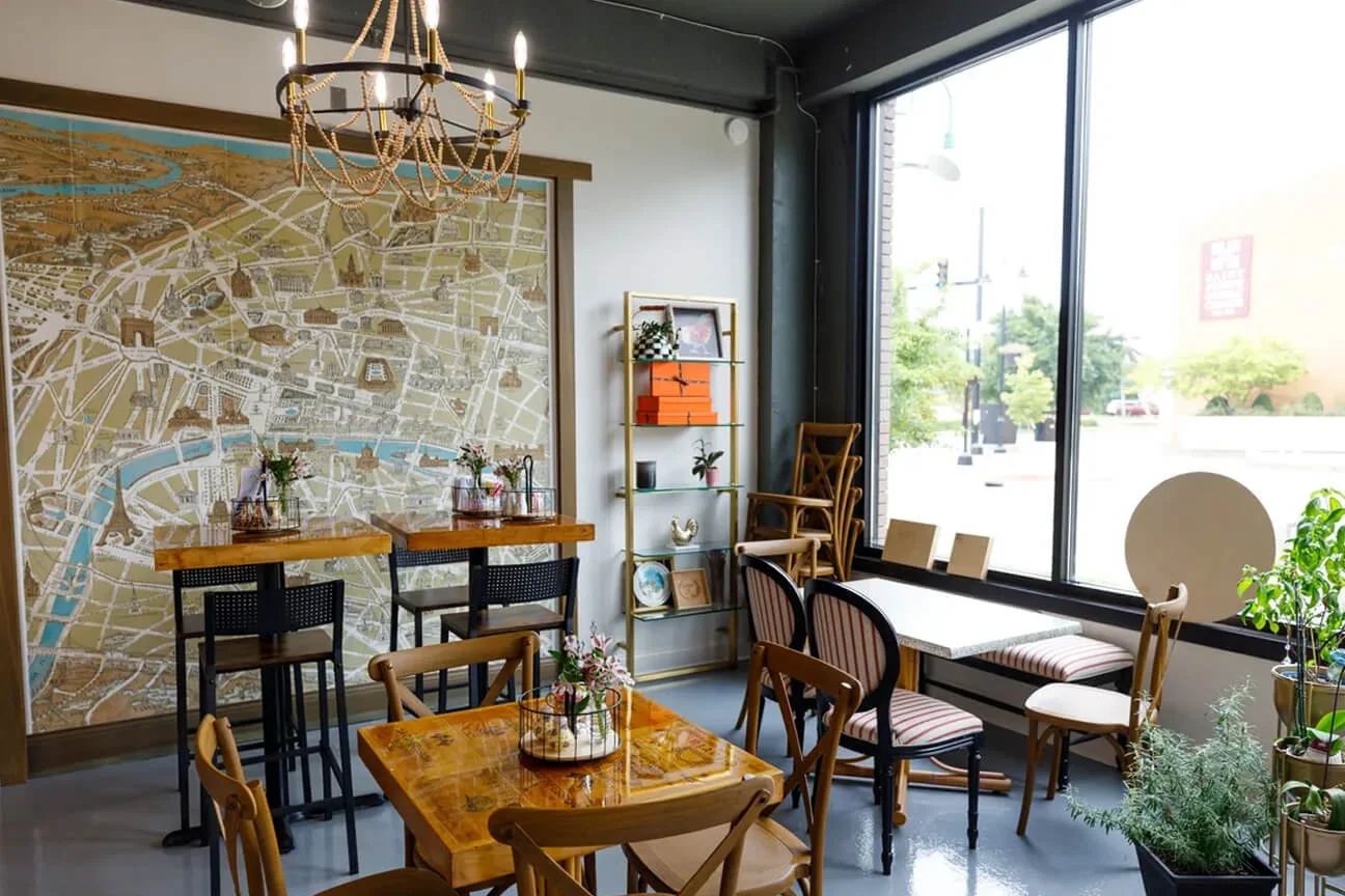 A cozy cafe corner with a large city map mural on the wall, wooden tables and chairs, a chandelier, and a large window letting in natural light. Decor includes plants and a shelf with books and decorative items.