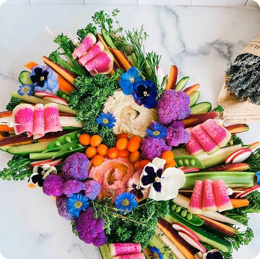 Colorful vegetable and flower bouquet arranged in a circular display on a white marble surface.