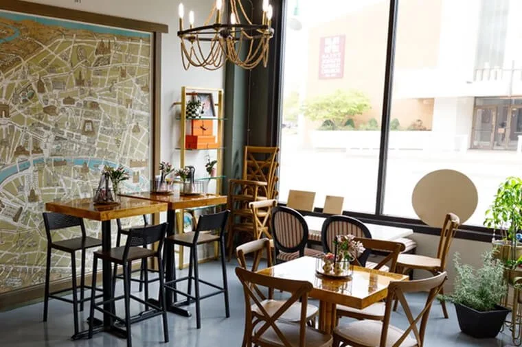 Interior of a cozy cafe with wooden tables and chairs, large windows, a map on the wall, and lush plants near the window.