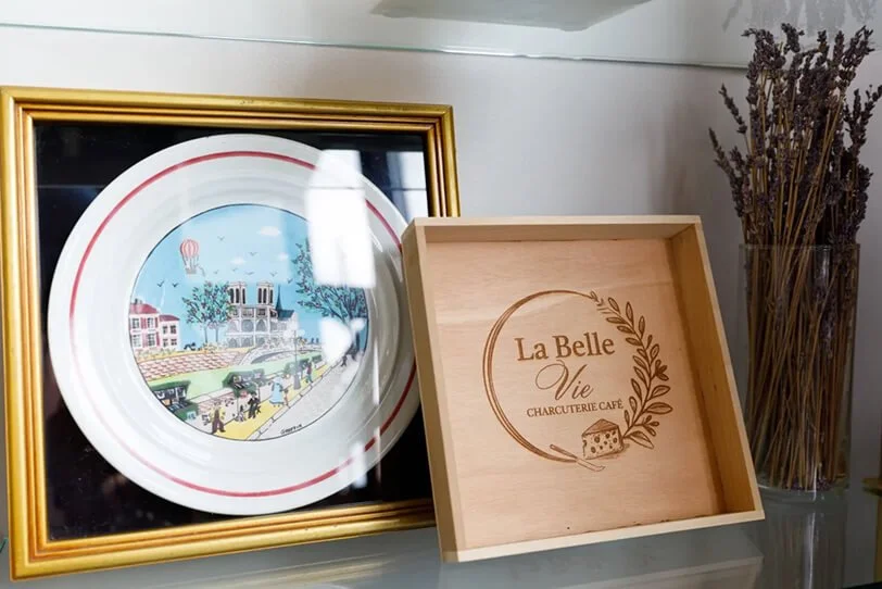 Decorative display with a framed plate featuring a colorful illustration of a cityscape, a wooden box with the words 'La Belle Vie' and 'Charcuterie Café', and a glass container filled with dried lavender branches.