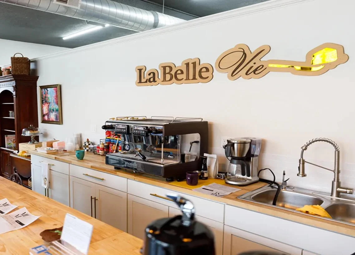 Coffee station with espresso machine, coffee maker, and sink, with a sign that says "La Belle Vie" on the wall.