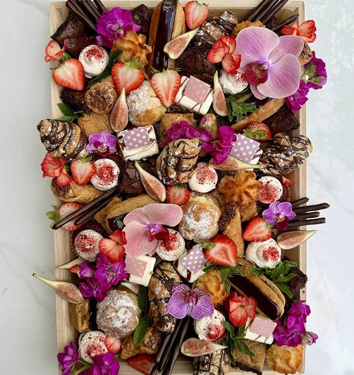 A large wooden box filled with an assortment of assorted desserts, strawberries, edible flowers, and decorative chocolates.