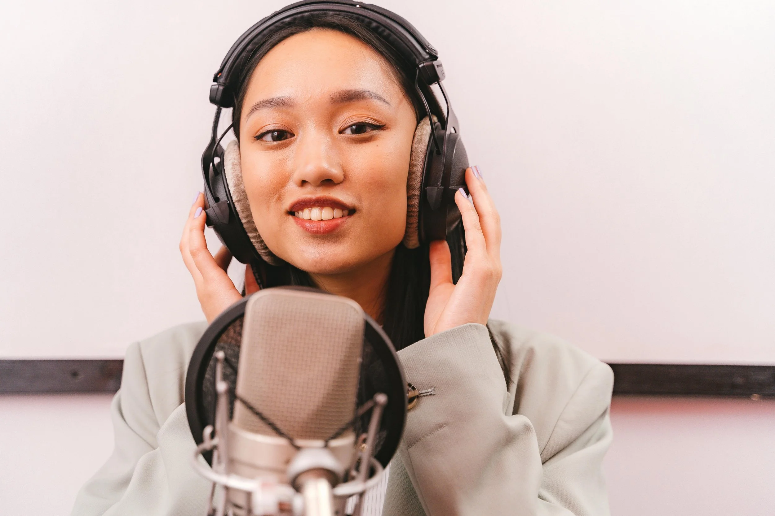 A young woman in a recording studio adjusting her headphones while standing behind a microphone.