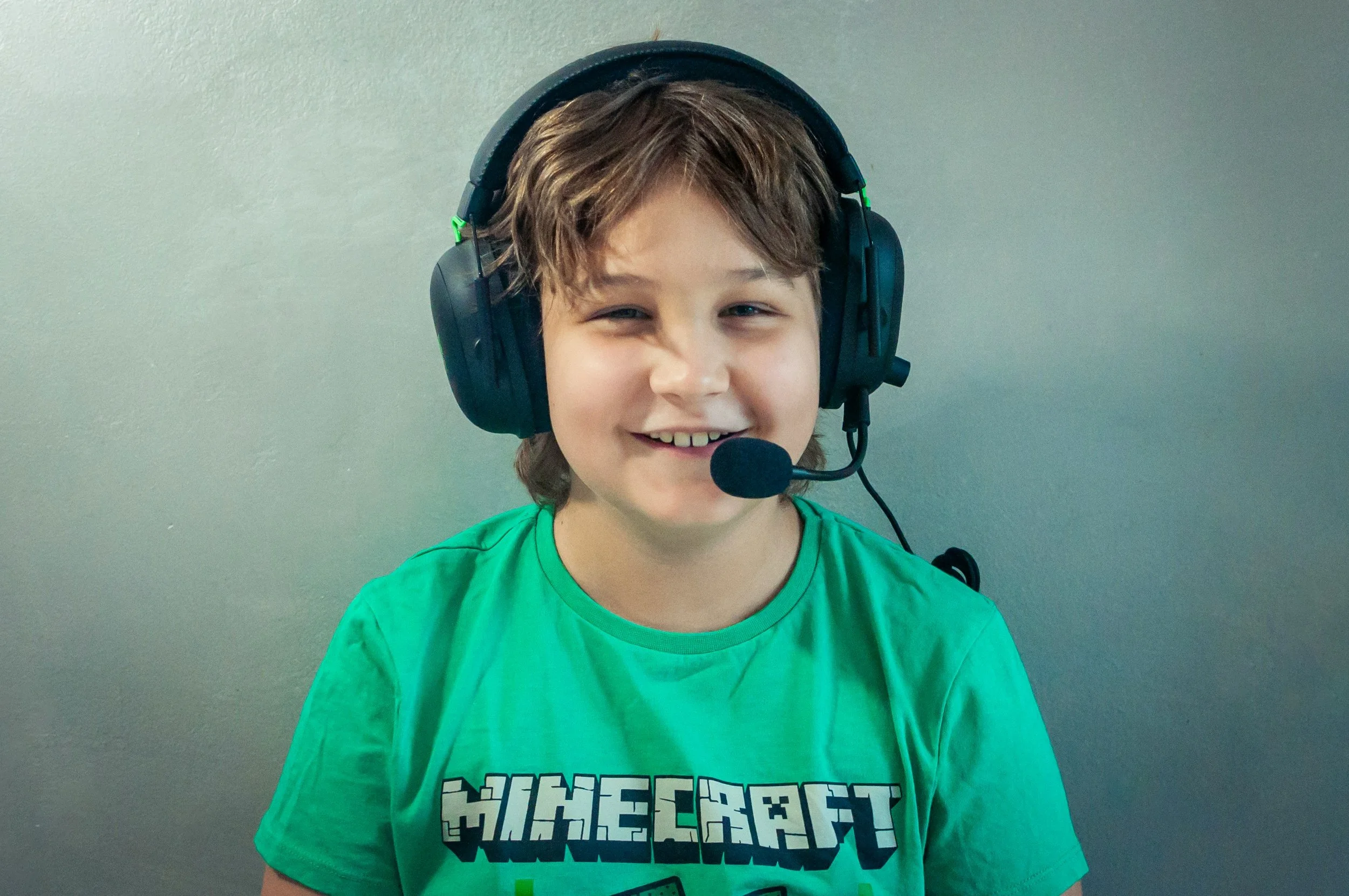 A young boy smiles while wearing a large black headset with a microphone, a green Minecraft-themed T-shirt, standing against a plain wall.
