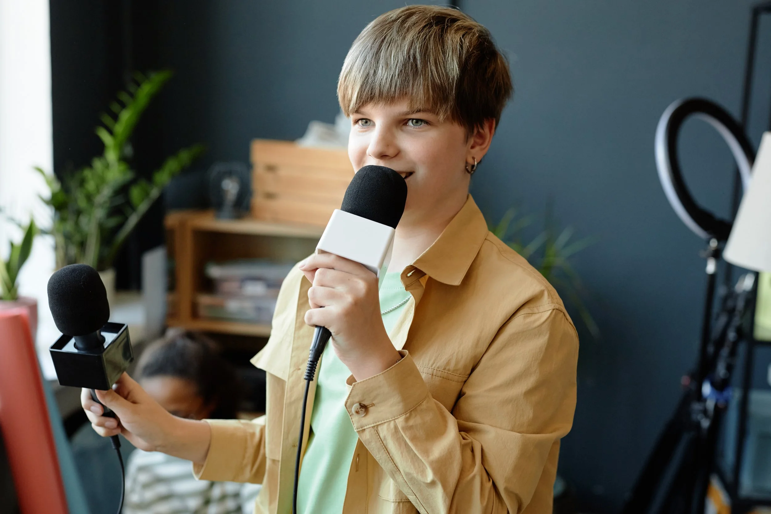 A young person with short, light brown hair and earrings is holding a microphone and speaking during a recording session. They are wearing a yellow jacket over a green shirt. There is a person in the background and indoor plants are visible on the left side of the image.