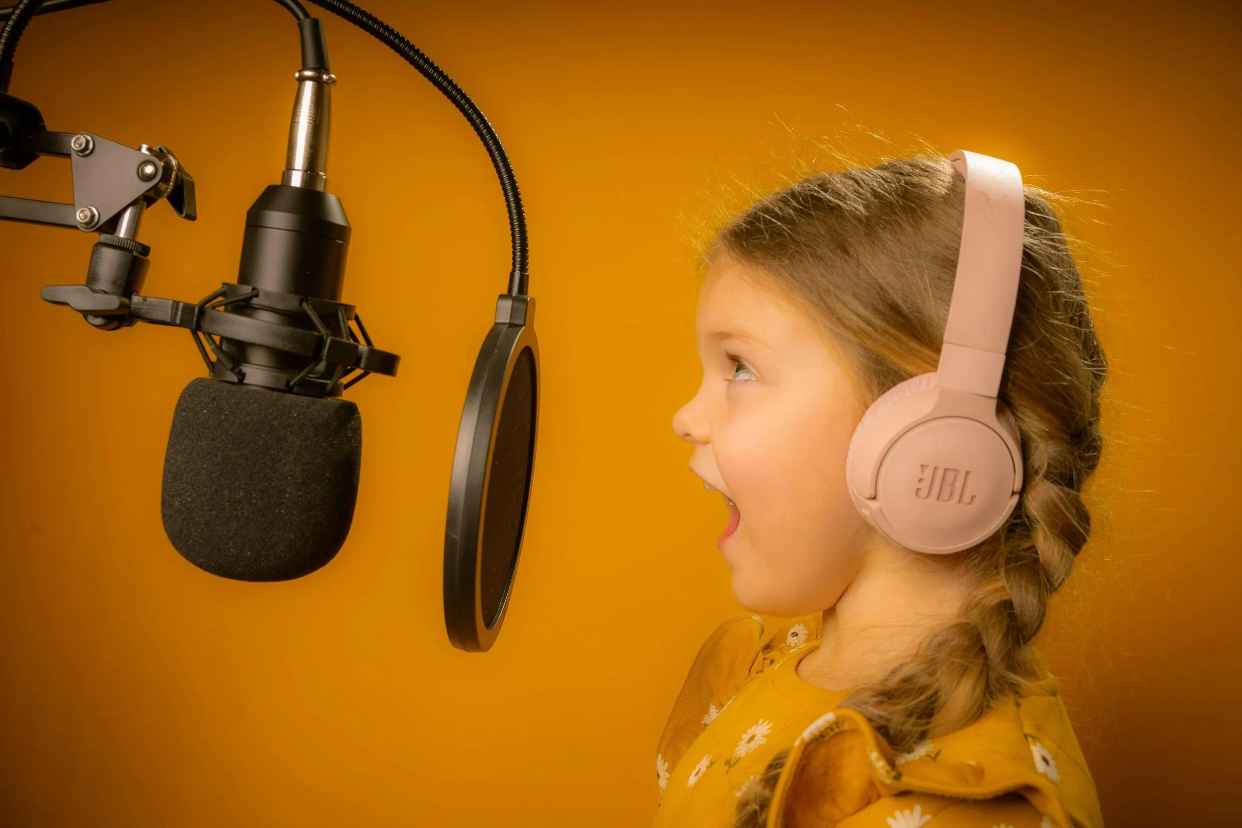 Young girl with braided hair singing into a music studio microphone while wearing pink headphones and a yellow dress with white flowers, against an orange background.