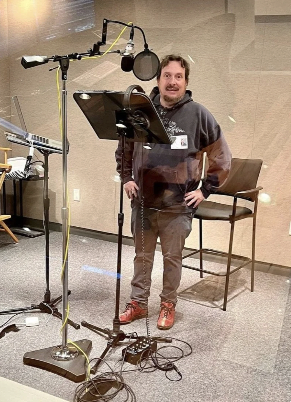 A man in casual clothing, standing behind a microphone and sound equipment in a recording studio, with a laptop on a stand and a chair nearby.
