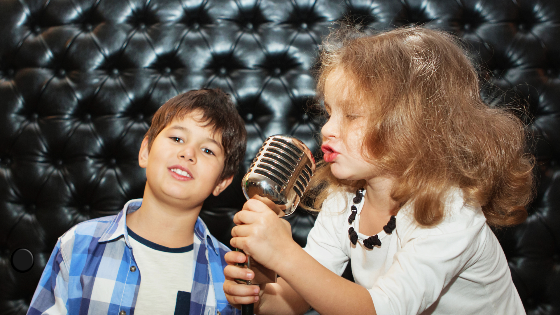 A young girl with curly red hair singing into a vintage-style microphone, while a young boy in a blue checkered shirt looks on, in front of a black tufted leather background.
