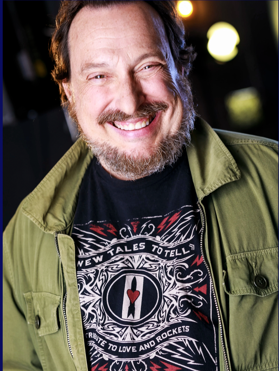 Smiling man with a beard and mustache, wearing a green jacket over a black graphic t-shirt with a heart and swirling design, standing outdoors at night with blurred lights in the background.