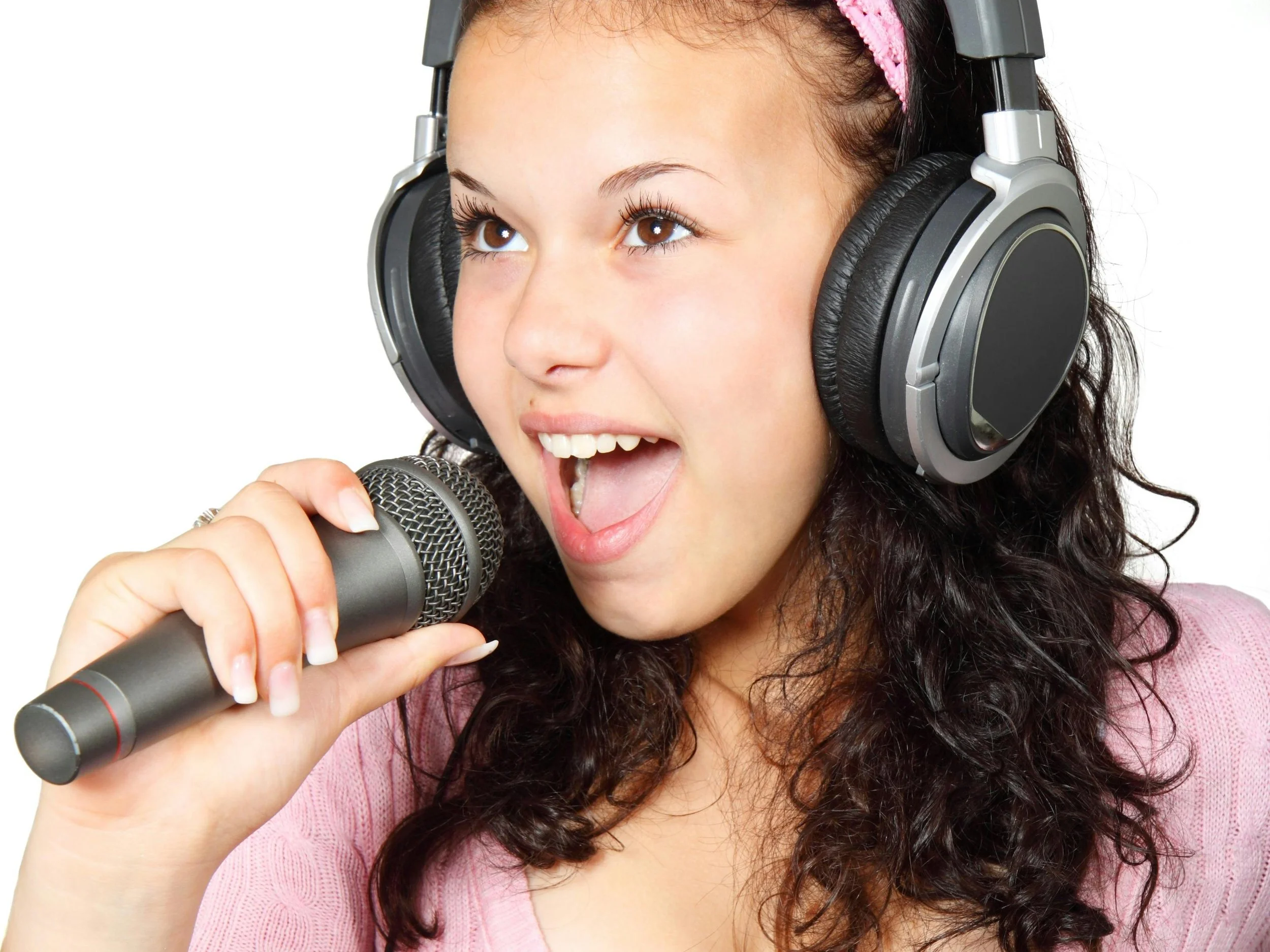 A young girl with dark curly hair wearing large black and gray headphones singing into a microphone, with an excited expression.