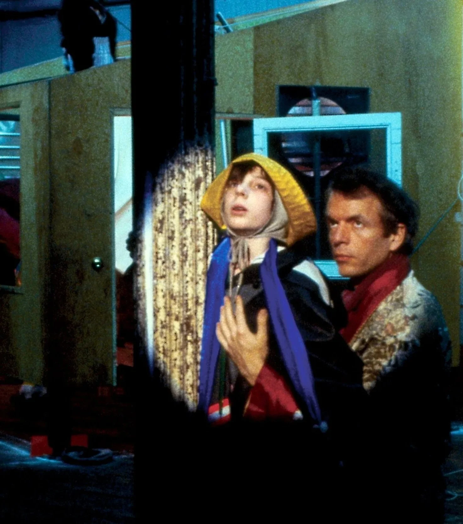 A young woman wearing a yellow hat and a beige headscarf, holding a coat, stands next to a man with dark hair and a patterned shirt. They are in a room with green walls, a black curtain, and various objects, including a television and bookshelves.