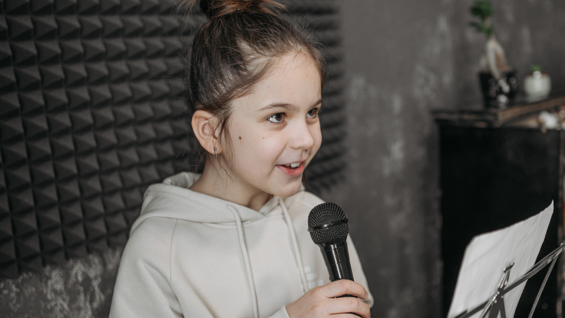 A young girl with brown hair tied in a bun, wearing a light-colored hoodie, is holding a black microphone and reading from a sheet of paper. She is sitting in front of soundproof foam panels.