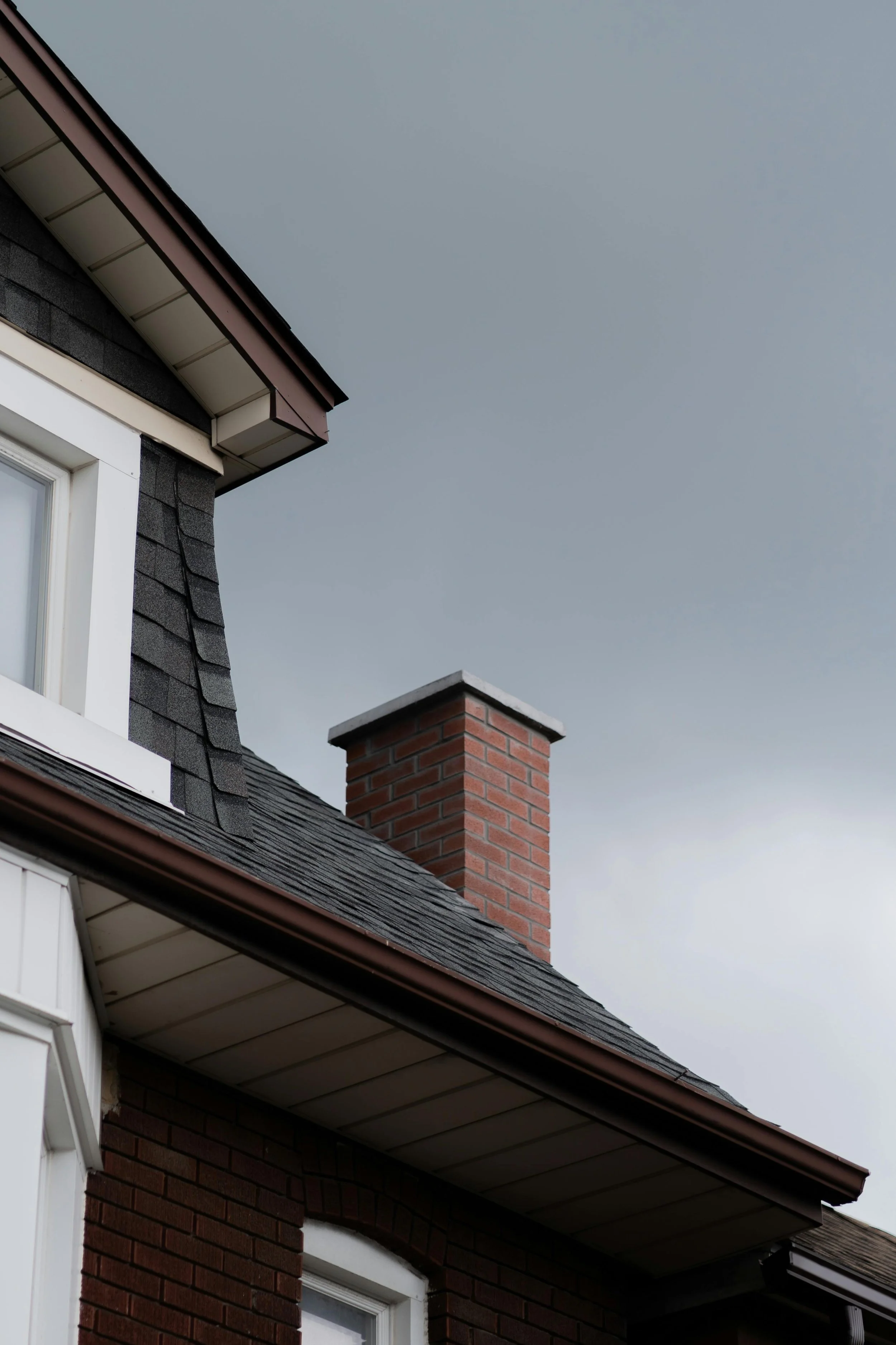 Close-up of a residential roofline with gray shingles, brick chimney, and white window trim.