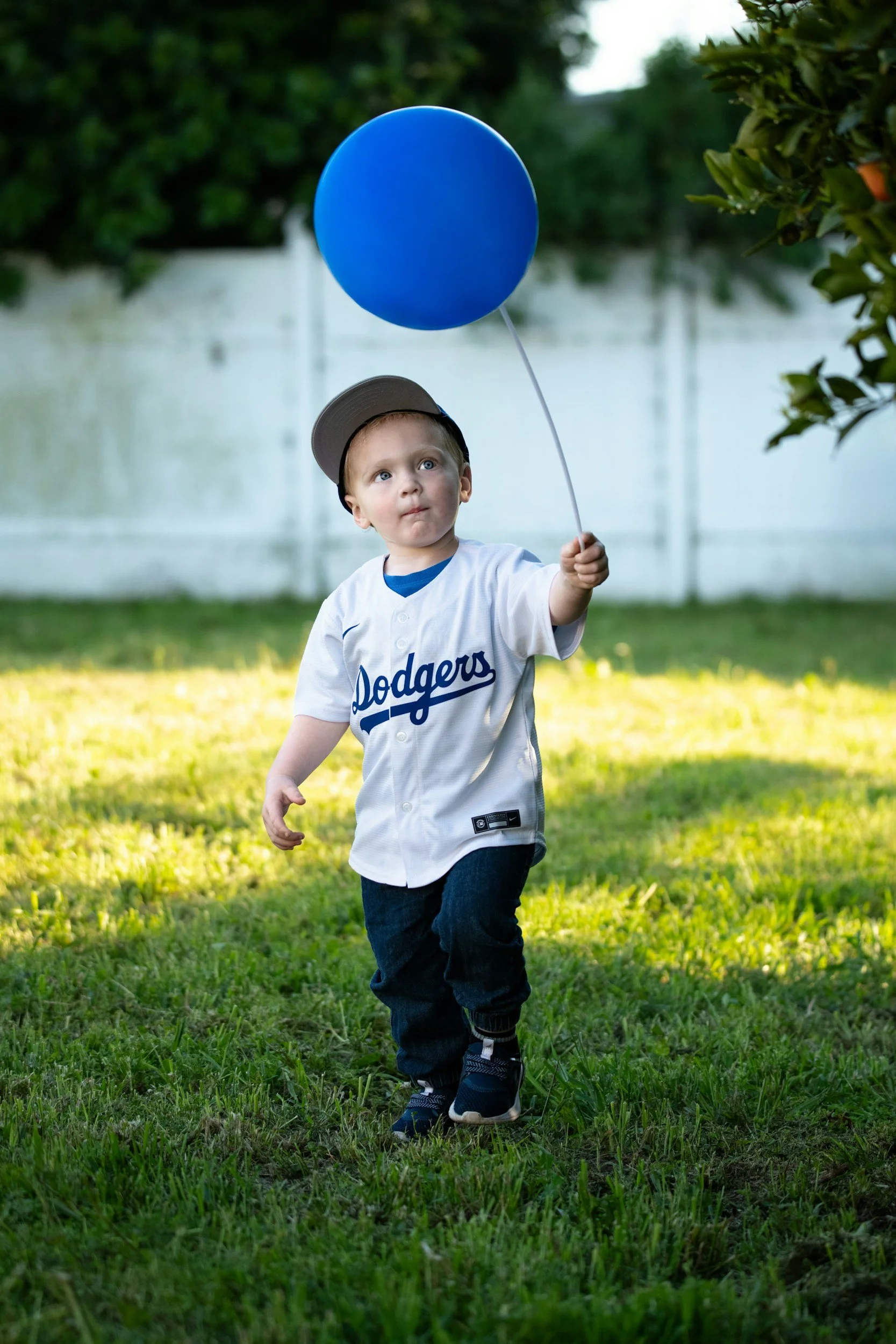 Young child in a Dodgers baseball jersey and cap walking on grass holding a blue balloon in an outdoor backyard setting.