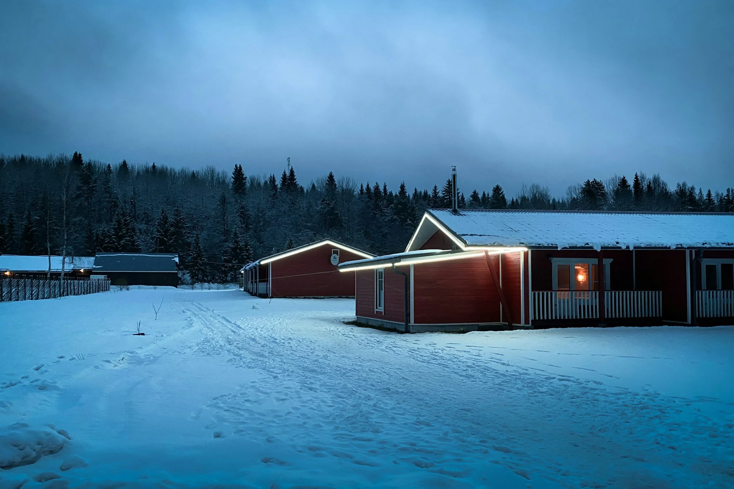 Snow-covered rural homes and trees at dusk with outdoor lights on.