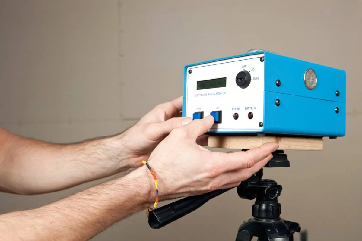 Person adjusting a blue and white continuous radiation monitor device mounted on a wooden platform on a tripod.