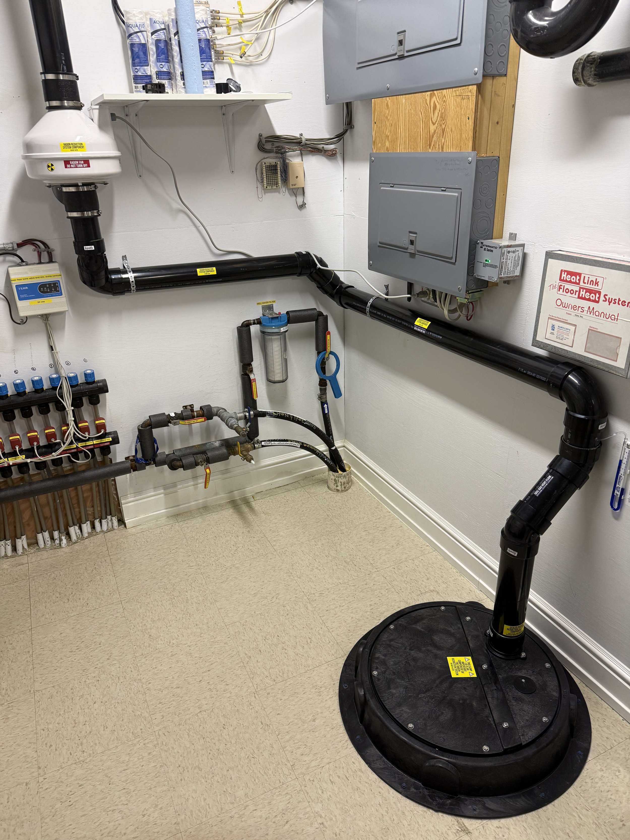 The image shows a mechanical room with plumbing and electrical panels. There is a large black round base connected to a vertical pipe, a white filter and piping, and various electrical boxes and wires on the wall.