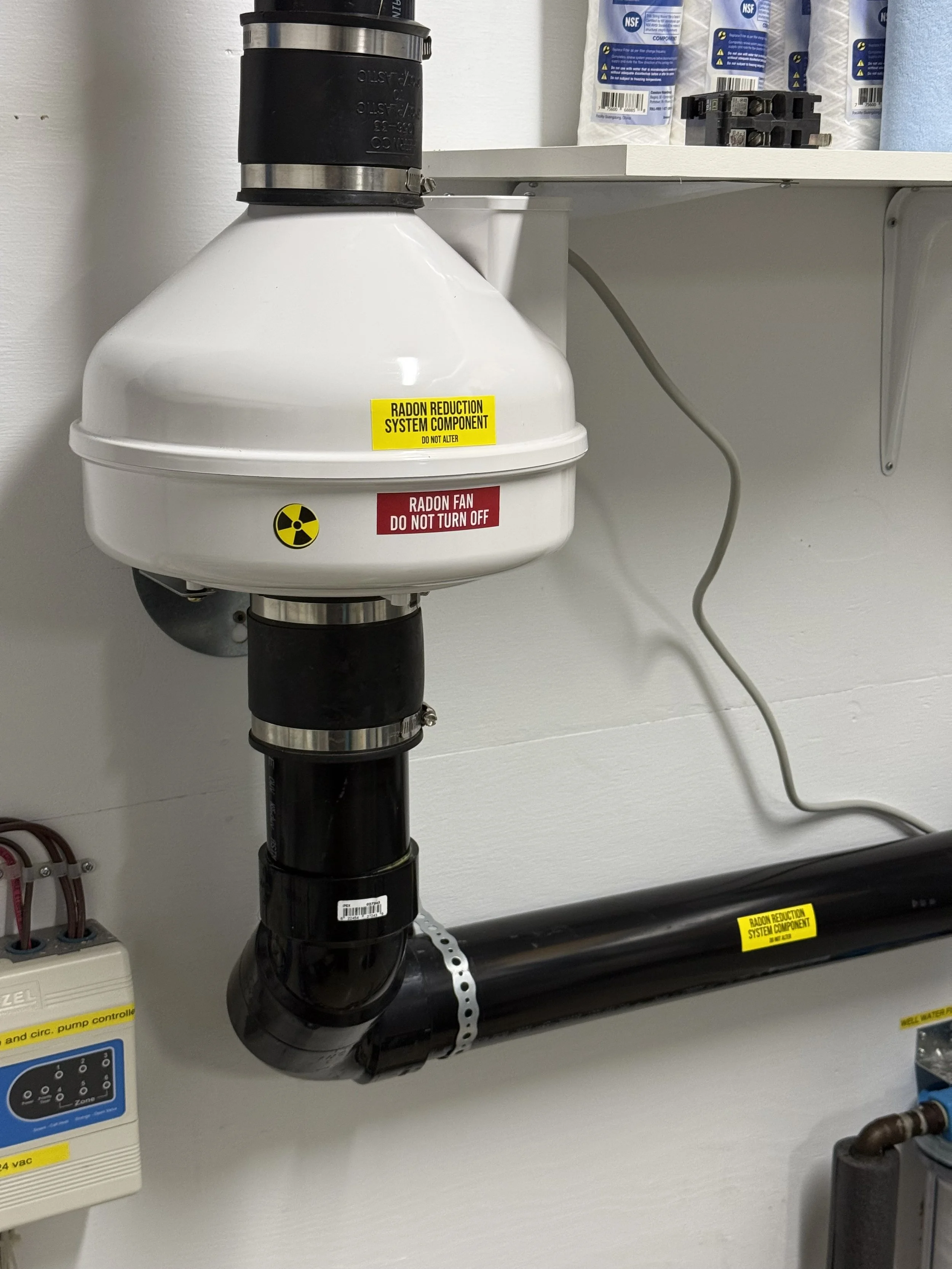 A white device with a yellow label that reads 'Radon Reduction System Component' connected to a black pipe system, with additional labels and controls nearby on a wall.