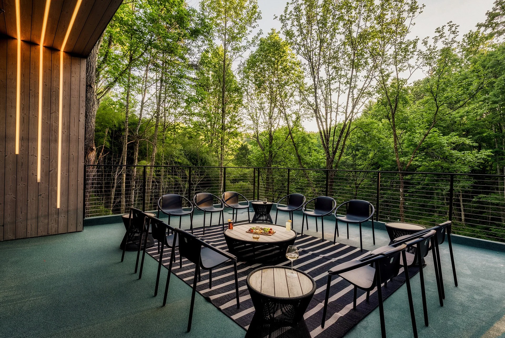 Outdoor balcony with black chairs and tables, surrounded by lush green trees and wooden wall with vertical lighting.