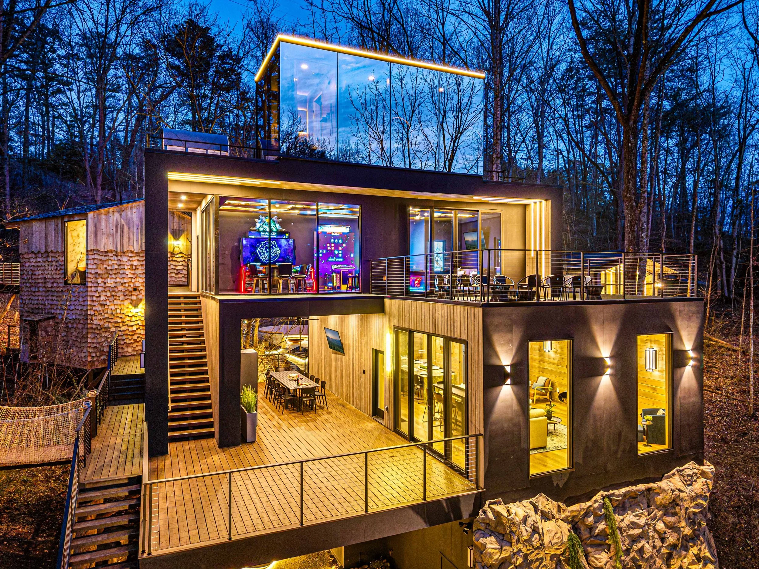 Modern multi-level house with glass walls and a balcony, illuminated at night, surrounded by leafless trees.