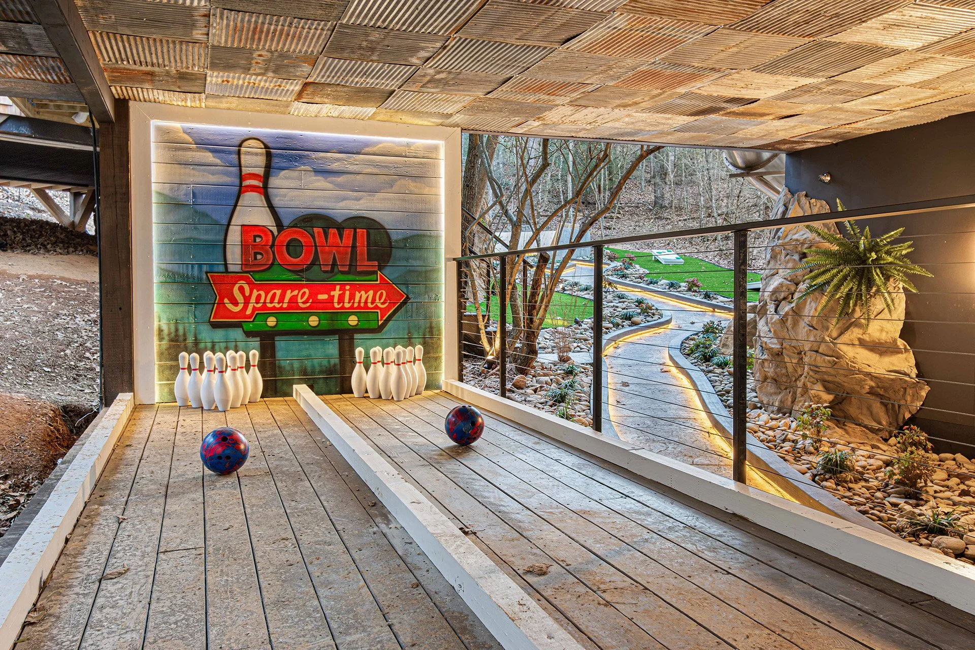 A mini bowling alley with a backdrop of a painted bowling icon with the words 'BOWL' and 'Spare-time' on a wooden wall. There are bowling balls and pins, with a landscaped outdoor area visible in the background.