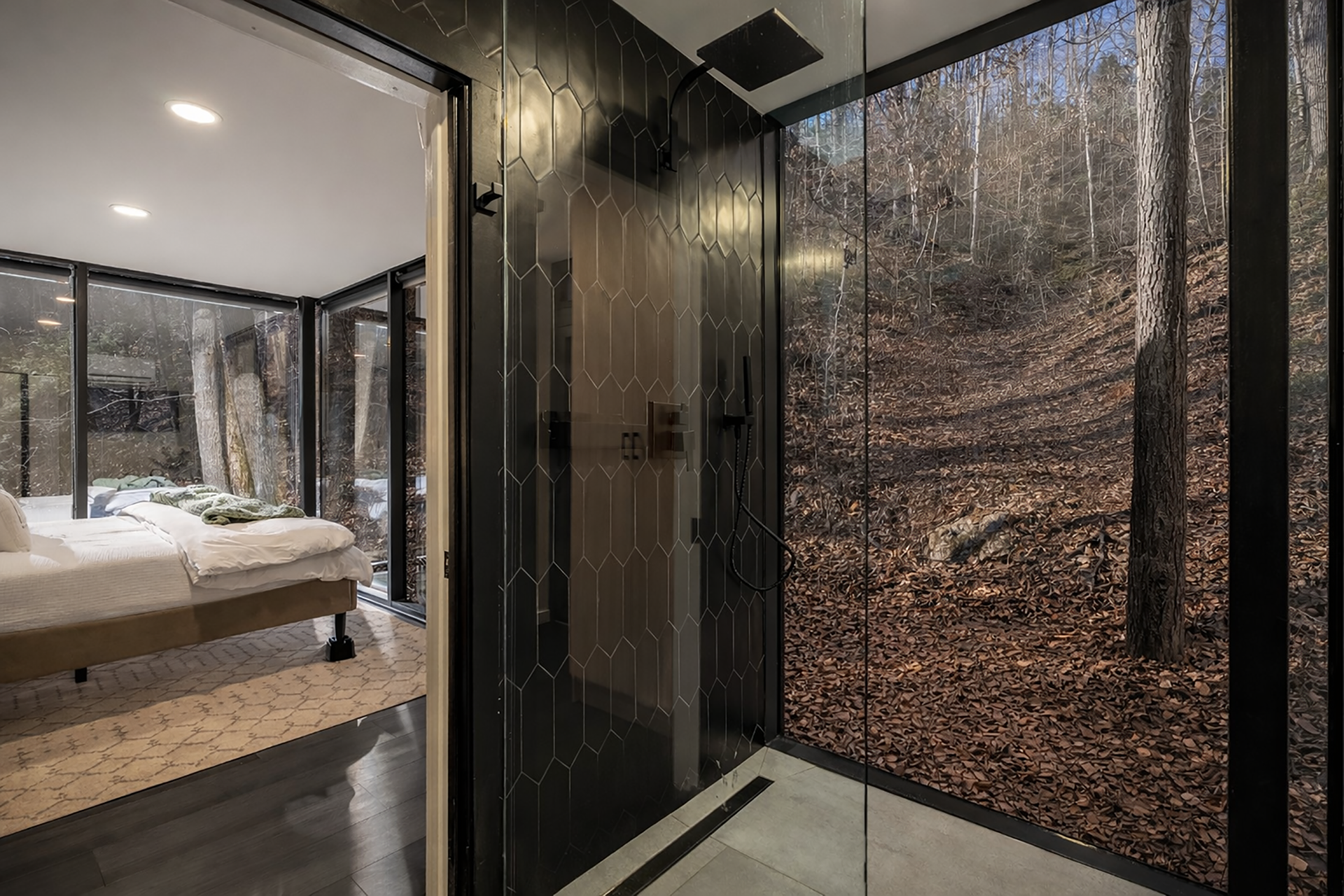 A modern bedroom with large windows showing trees outside, beside a bathroom with a digital shower panel and a wall mural of a forest scene.