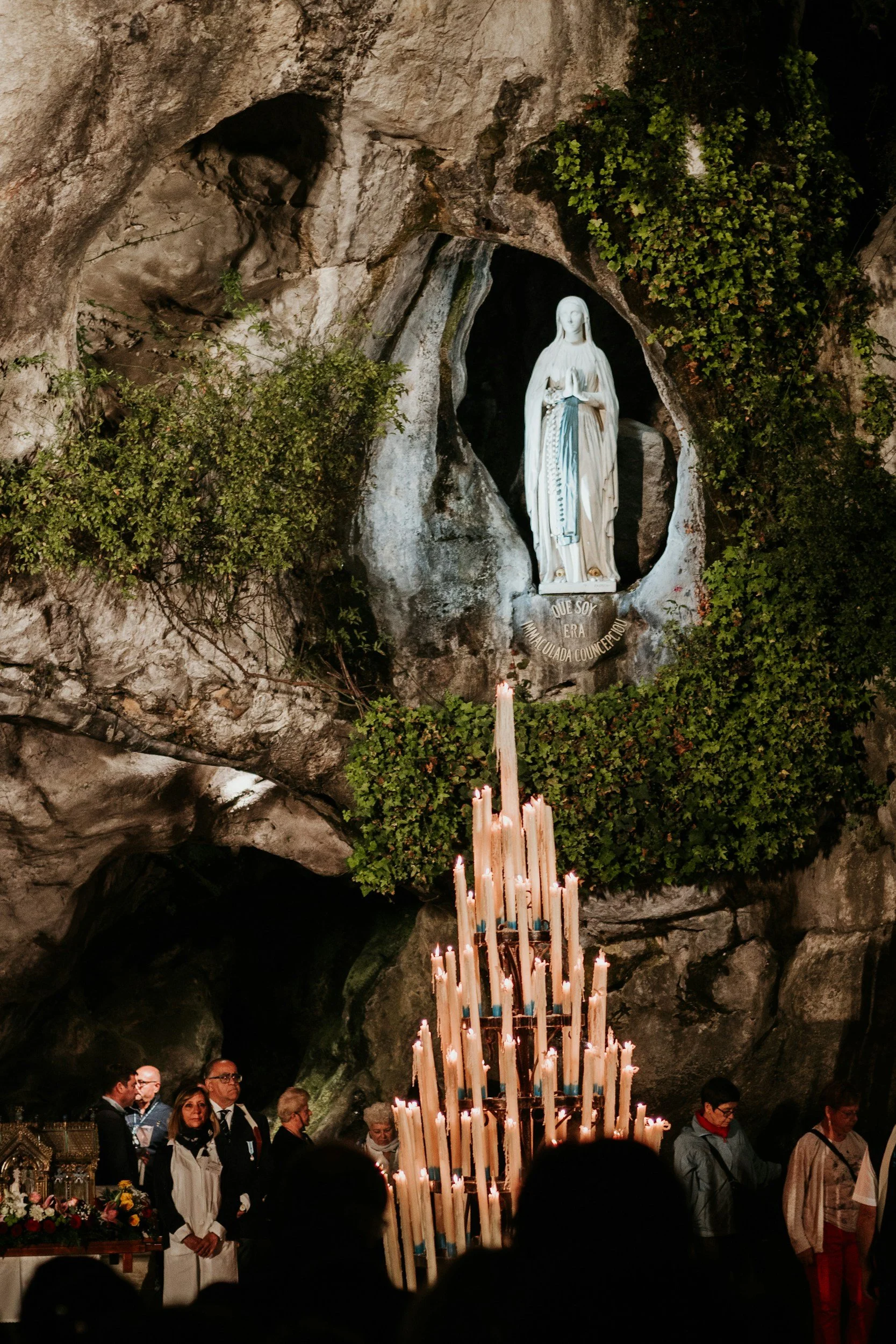 People gathered inside a cave with a religious statue of the Virgin Mary at the altar. The altar is adorned with lit candles, and there are plants and flowers around. The statue is positioned within a natural rock formation, and some individuals are seen praying or observing.