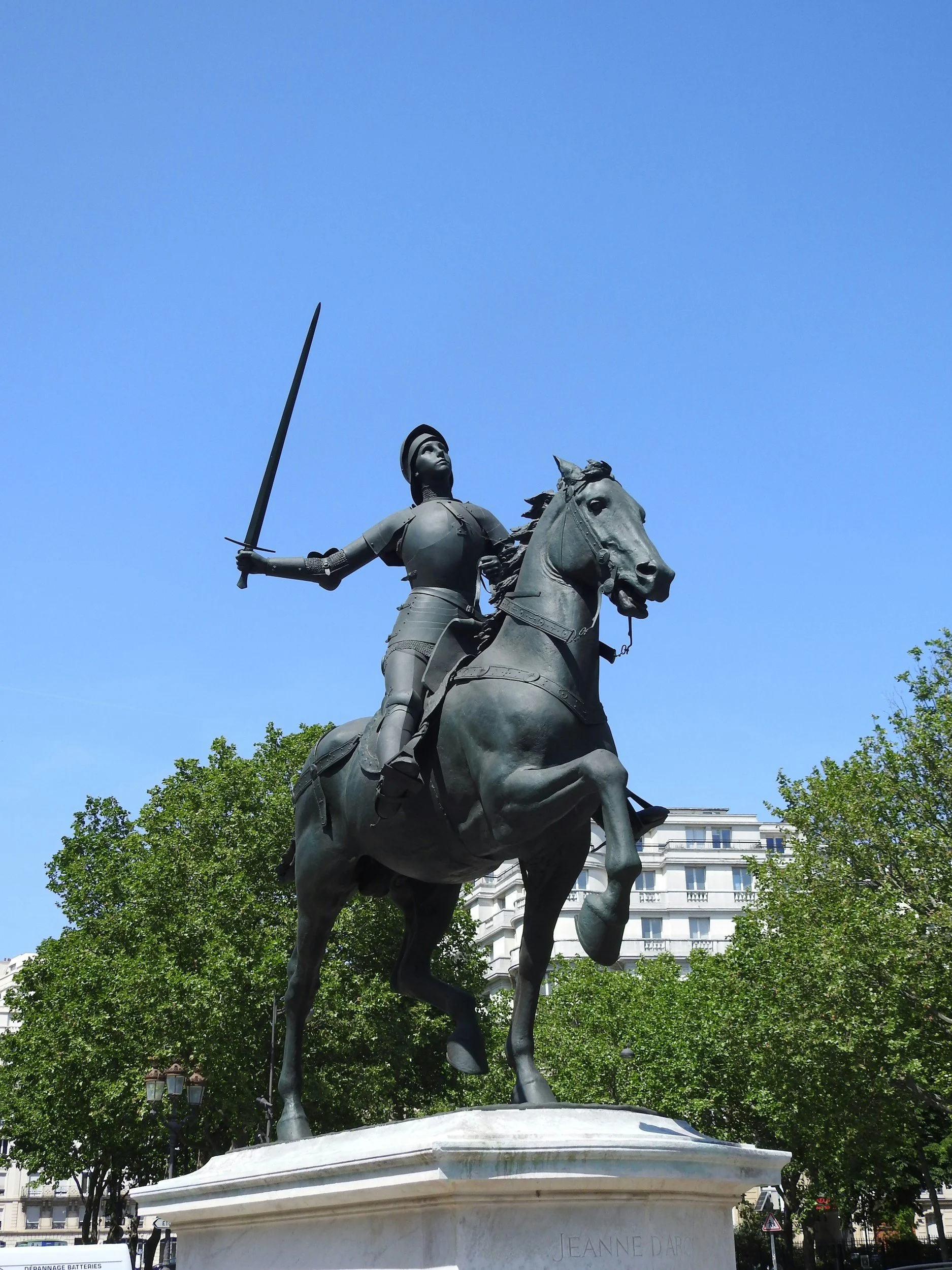 Brass equestrian statue of Jeanne d'Arc (Joan of Arc) on a white stone pedestal, set against a background of trees and a building, under a clear blue sky.