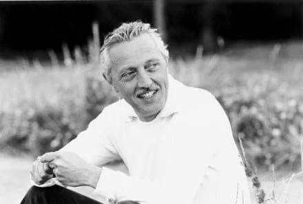 A black and white photo of a smiling man with light hair, sitting outdoors in a field, wearing a light-colored shirt.