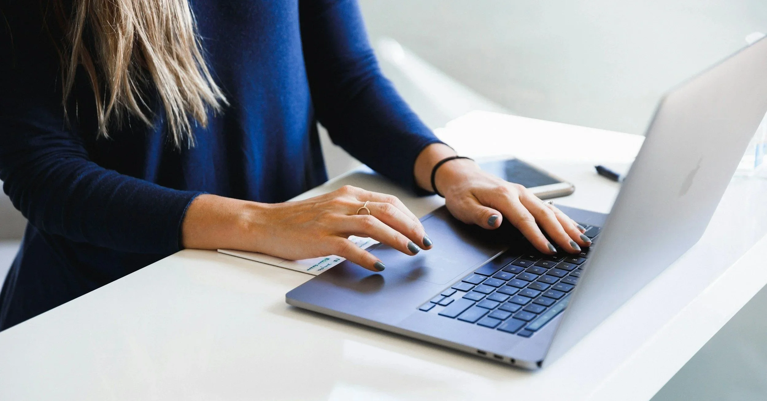 A person typing on a laptop at a white table, with a smartphone and notepad nearby.