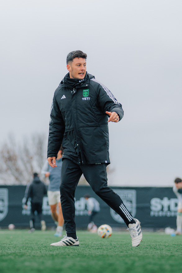A man in a black sports jacket and black pants, pointing and yelling, on a soccer field during training or a match.