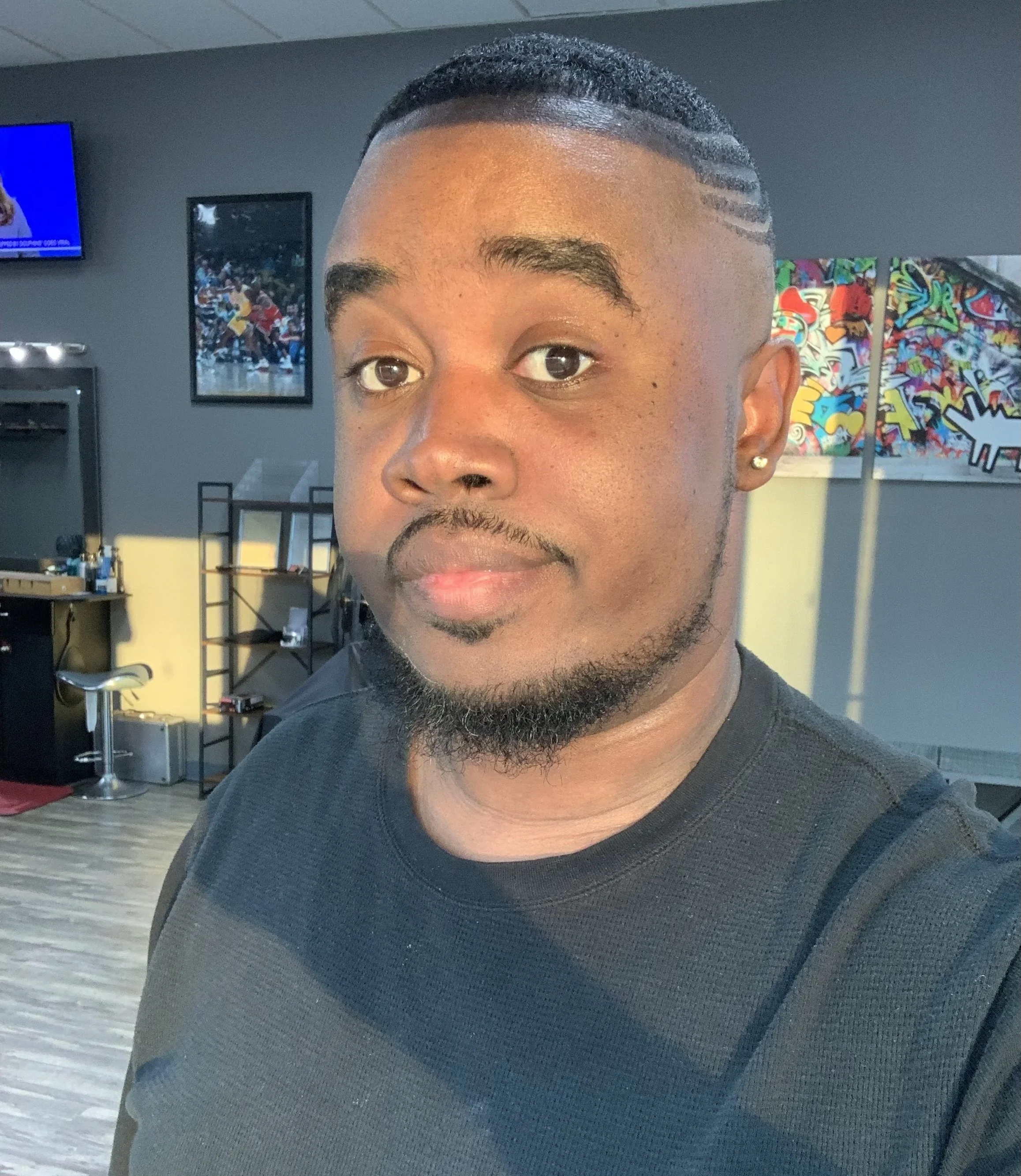 A young man with a short haircut, beard, and earrings, taking a selfie indoors with sunlight on his face.