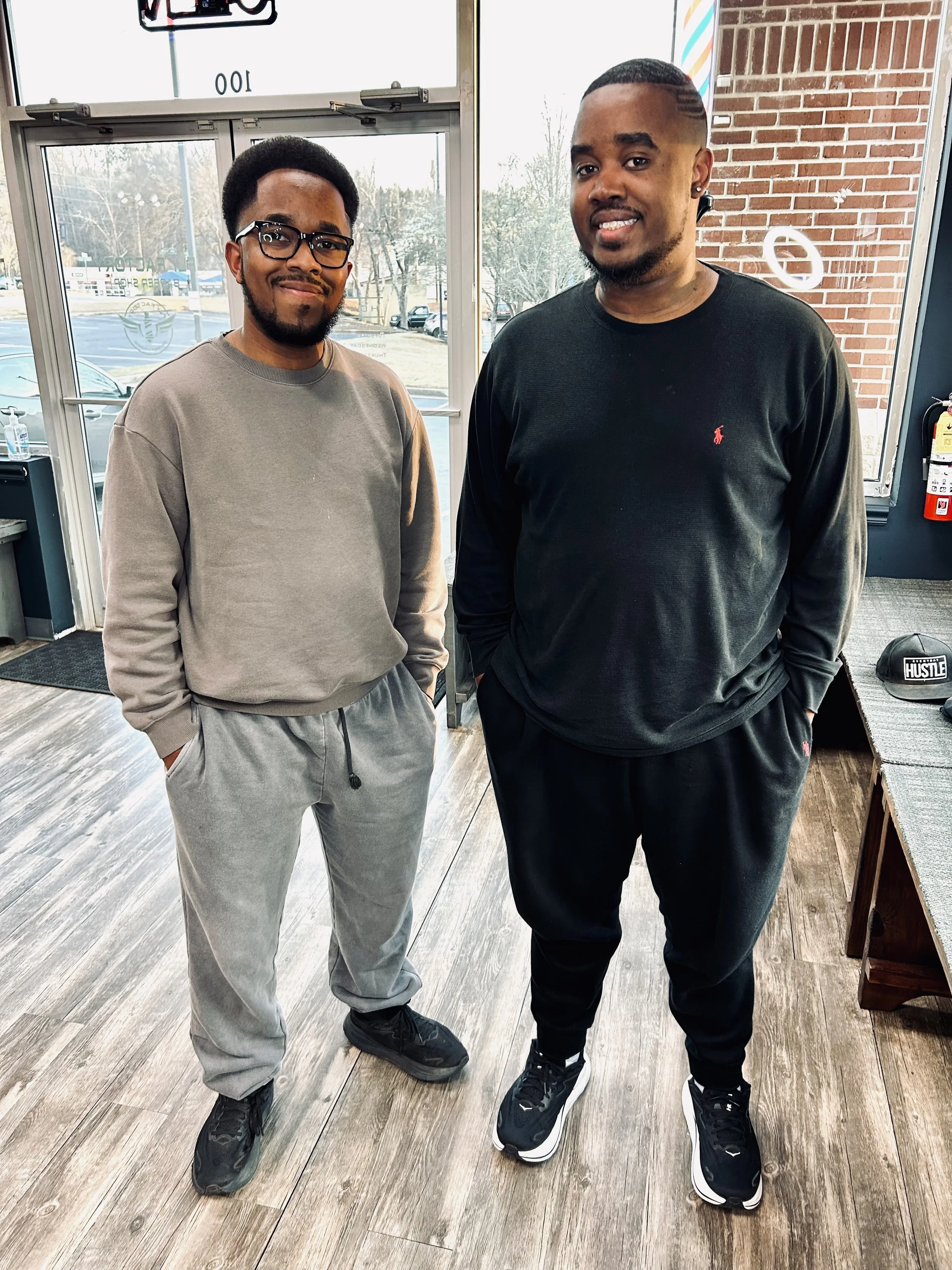 Two men standing inside a store, both smiling and dressed casually in sweatshirts and joggers, with a brick wall and large glass doors behind them.
