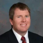 A headshot of a man with short brown hair, wearing a dark suit jacket, white shirt, and a red tie, smiling.