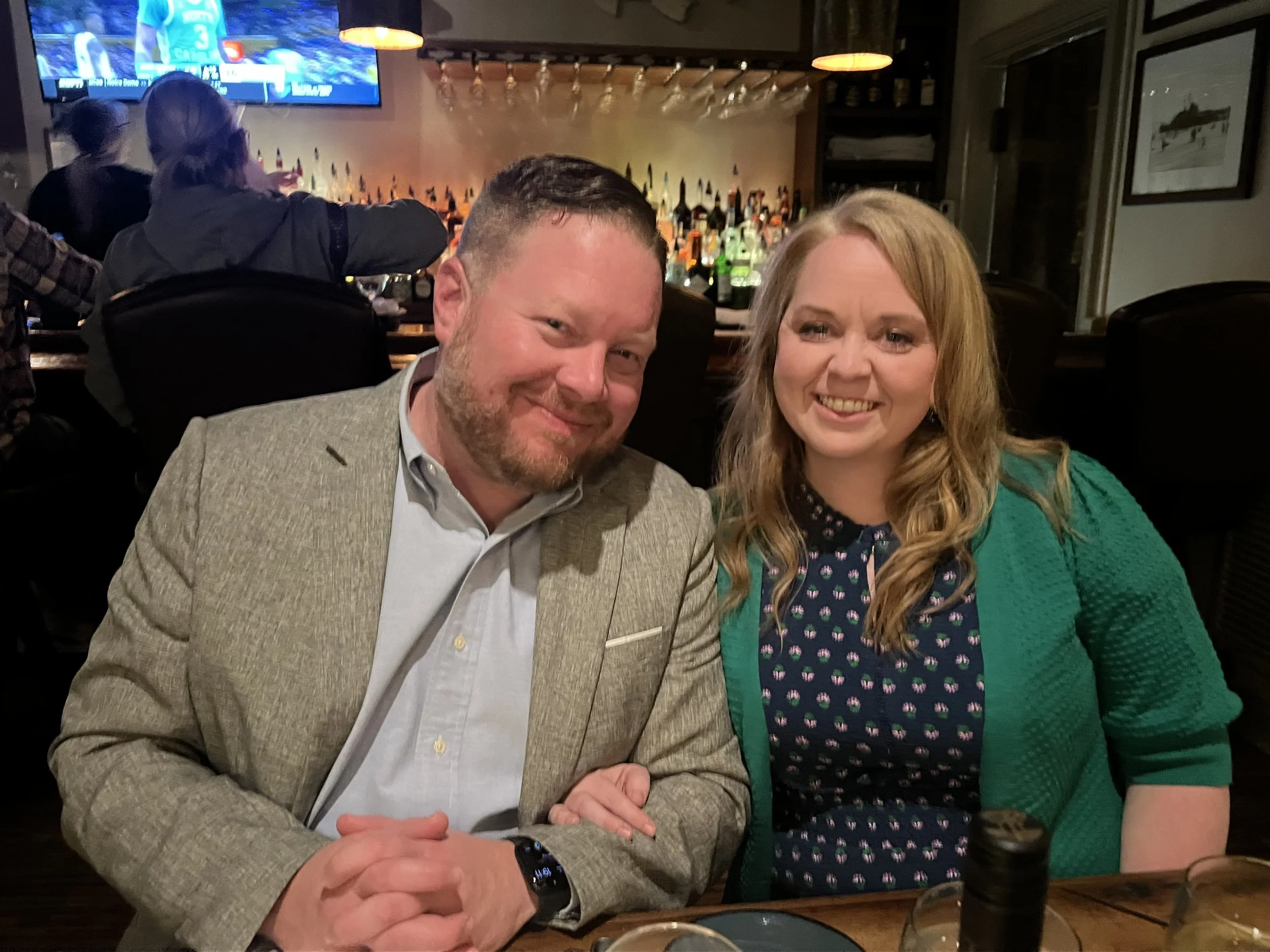 A man and woman sitting together at a restaurant bar, smiling at the camera. The man is wearing a light gray blazer and white shirt, and has a beard. The woman has long, wavy blonde hair, and is wearing a green cardigan over a dark dress with a pattern. The background shows a bar with bottles and glasses, and TVs mounted on the wall.