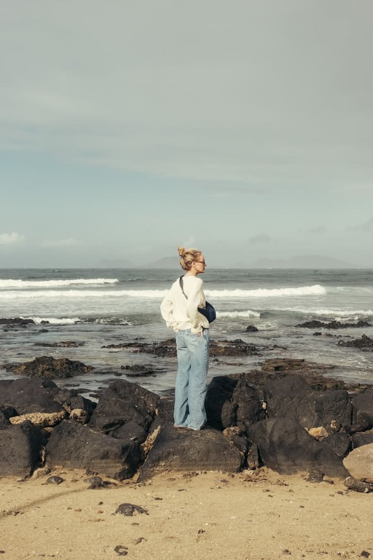 Woman standing on rocks at the beach, facing the ocean with waves, under cloudy sky.