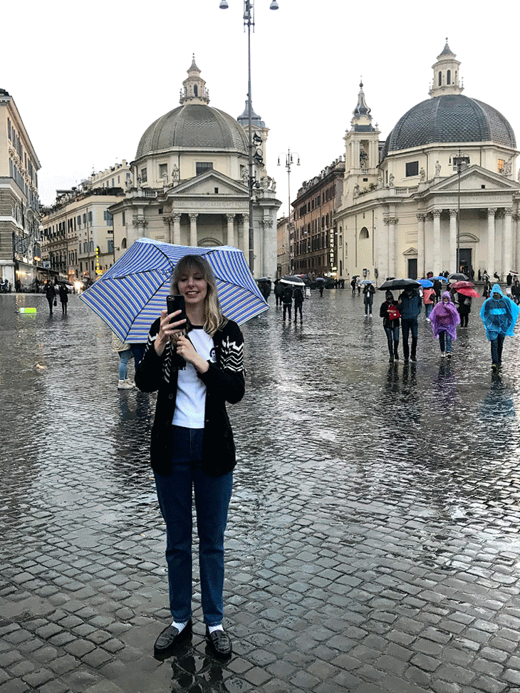 A woman smiling and taking a selfie with a smartphone on a rainy day in a European city square with historic buildings and church domes in the background. She is holding a blue striped umbrella, and people with umbrellas are walking by on wet cobblestone streets.