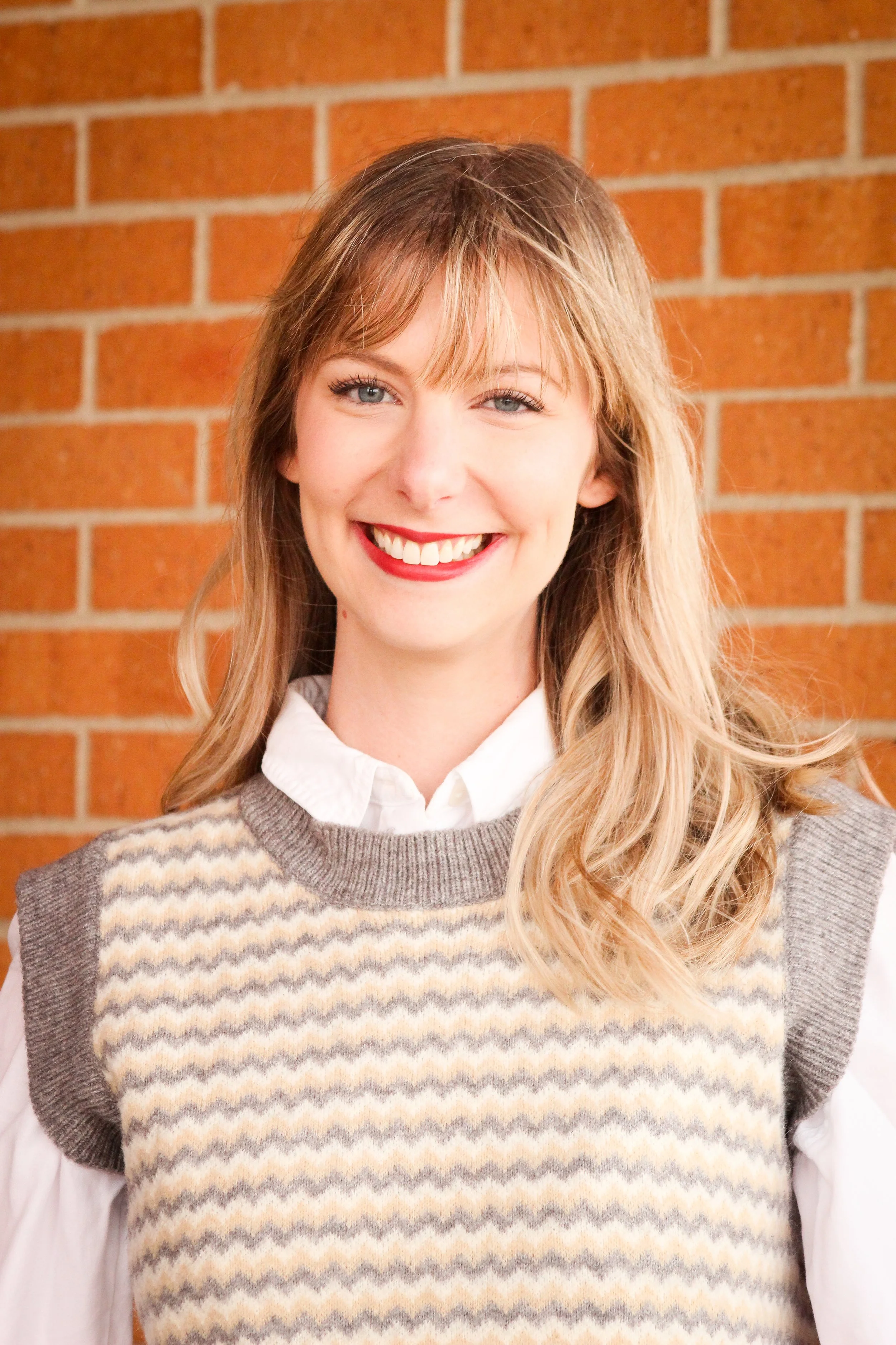 A woman with long blonde hair wearing a white shirt and a knitted sweater vest, smiling in front of a brick wall.