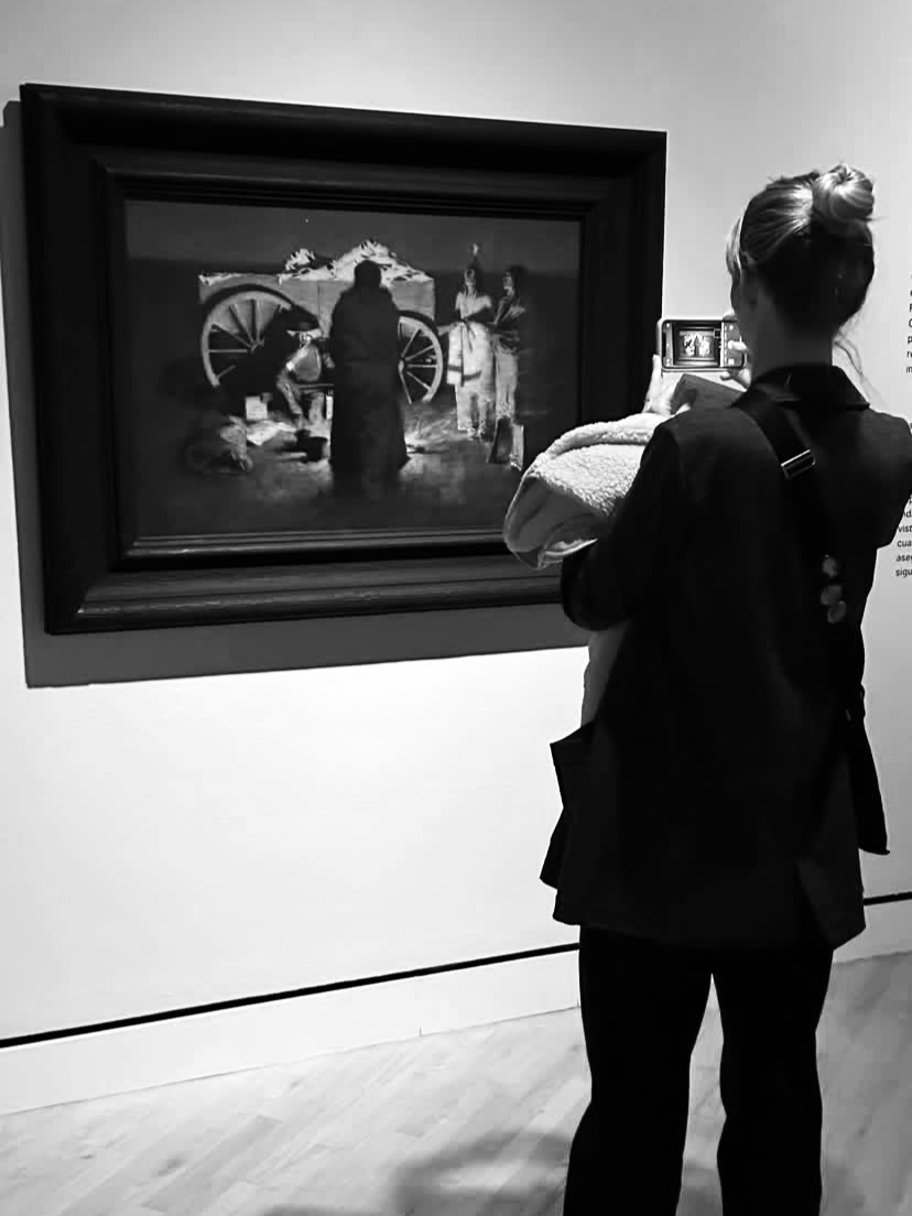 A woman with a bun hairstyle wearing a blazer is taking a photo of a painting in an art gallery.