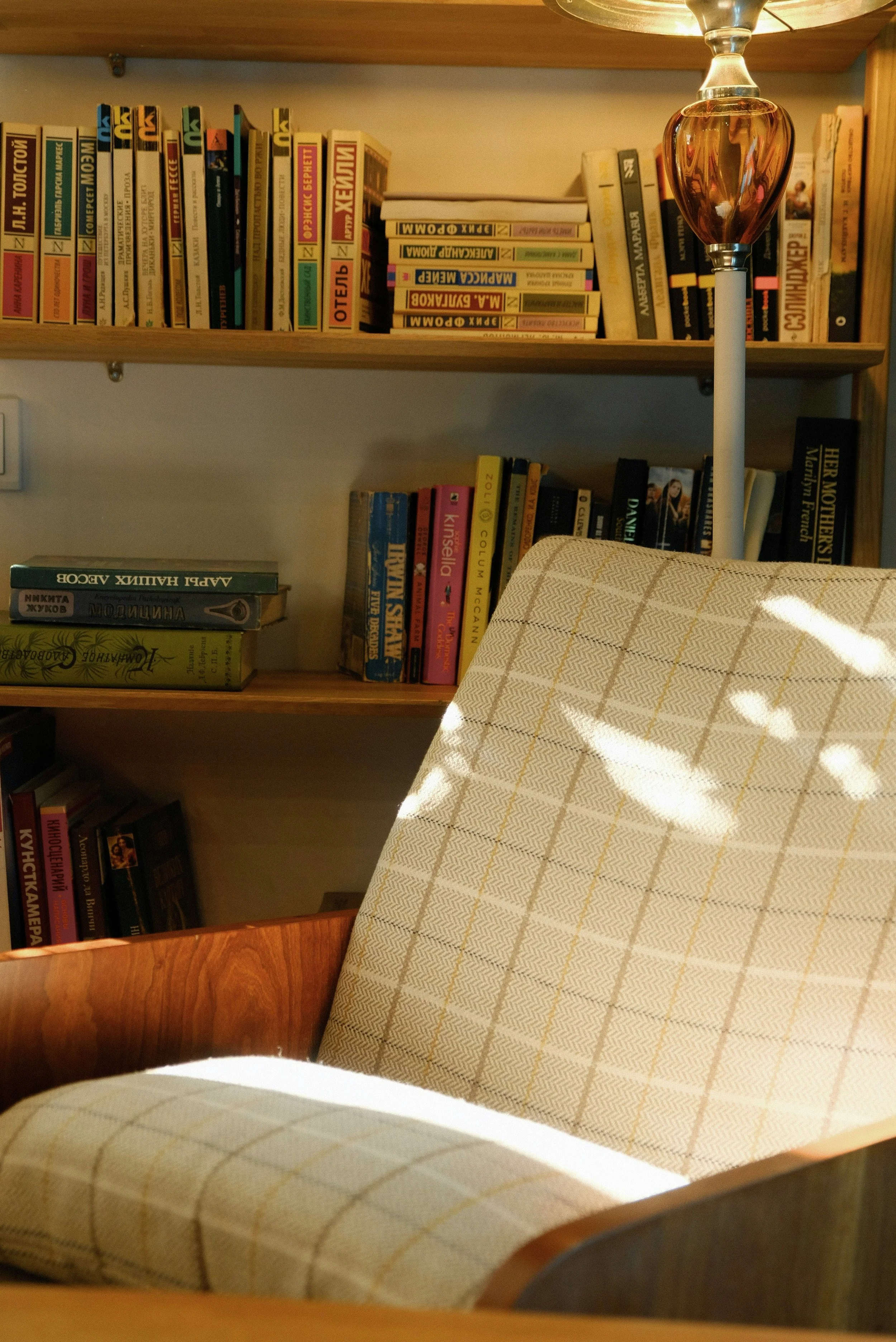 A cozy room with a bookshelf filled with books, a lamp with a pinkish glass shade, and a beige armchair with a plaid pattern. Sunlight creates shadows on the chair and wall.