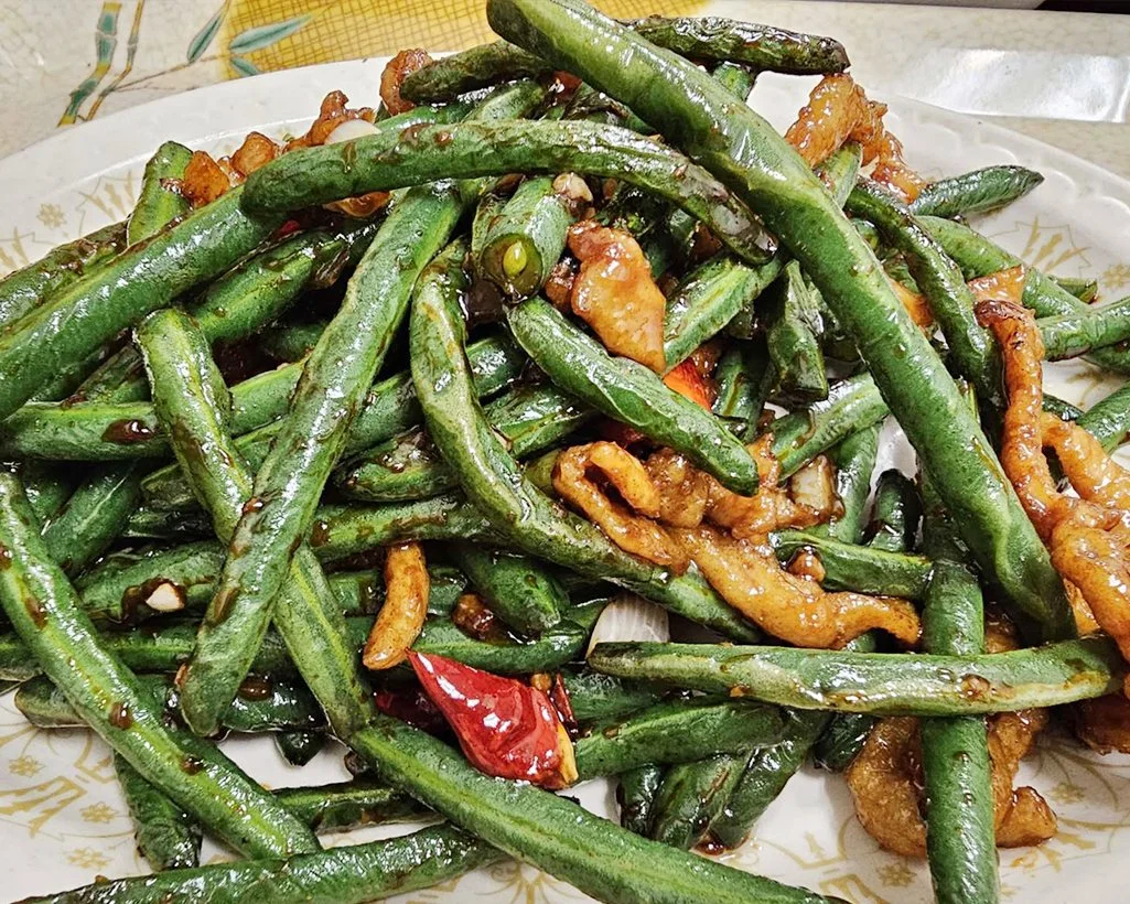 Stir-fried green beans with pieces of fried chicken, red chili peppers, and garlic, served on a white plate.