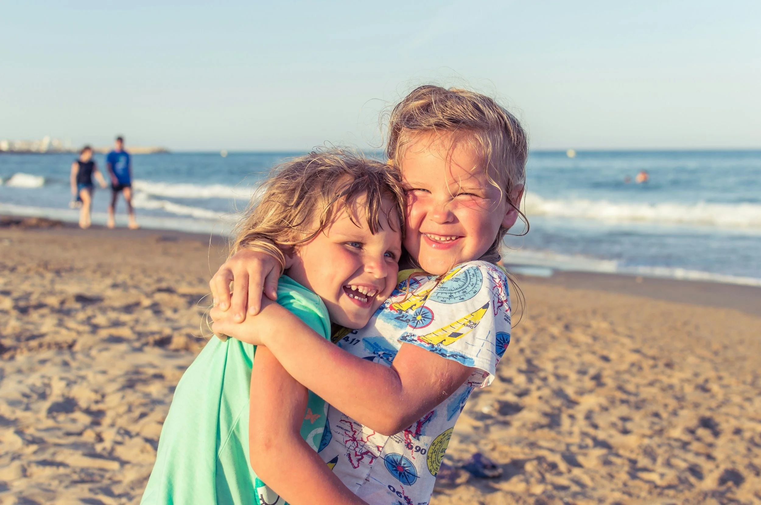 Two young children, a girl and a boy, hugging happily on a sandy beach with ocean waves and people swimming in the background. Education is not just schooling.