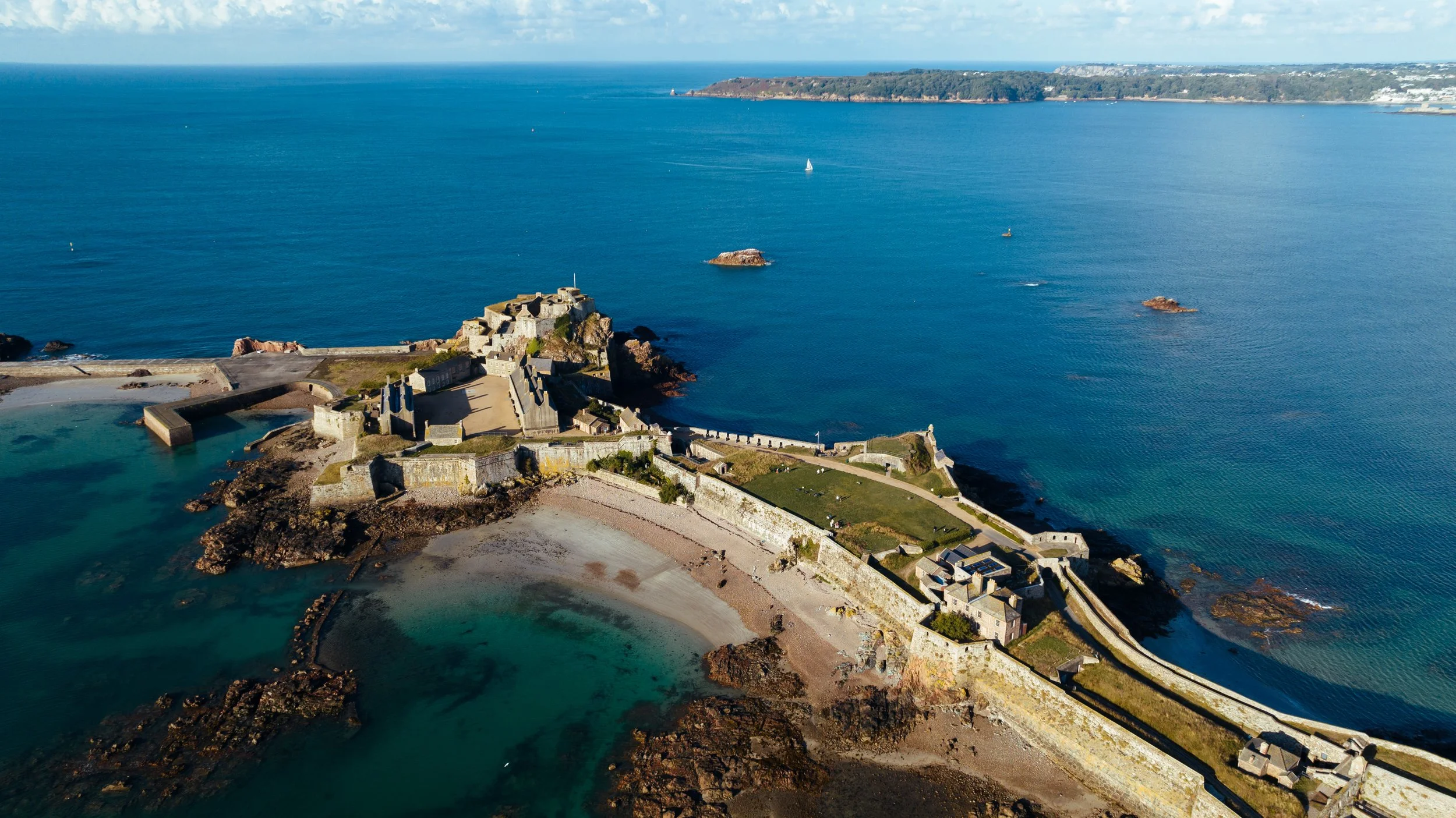 Aerial view of a historic coastal fortress on a rocky peninsula with walls, buildings, and a small harbor, surrounded by blue ocean water and a distant landmass.