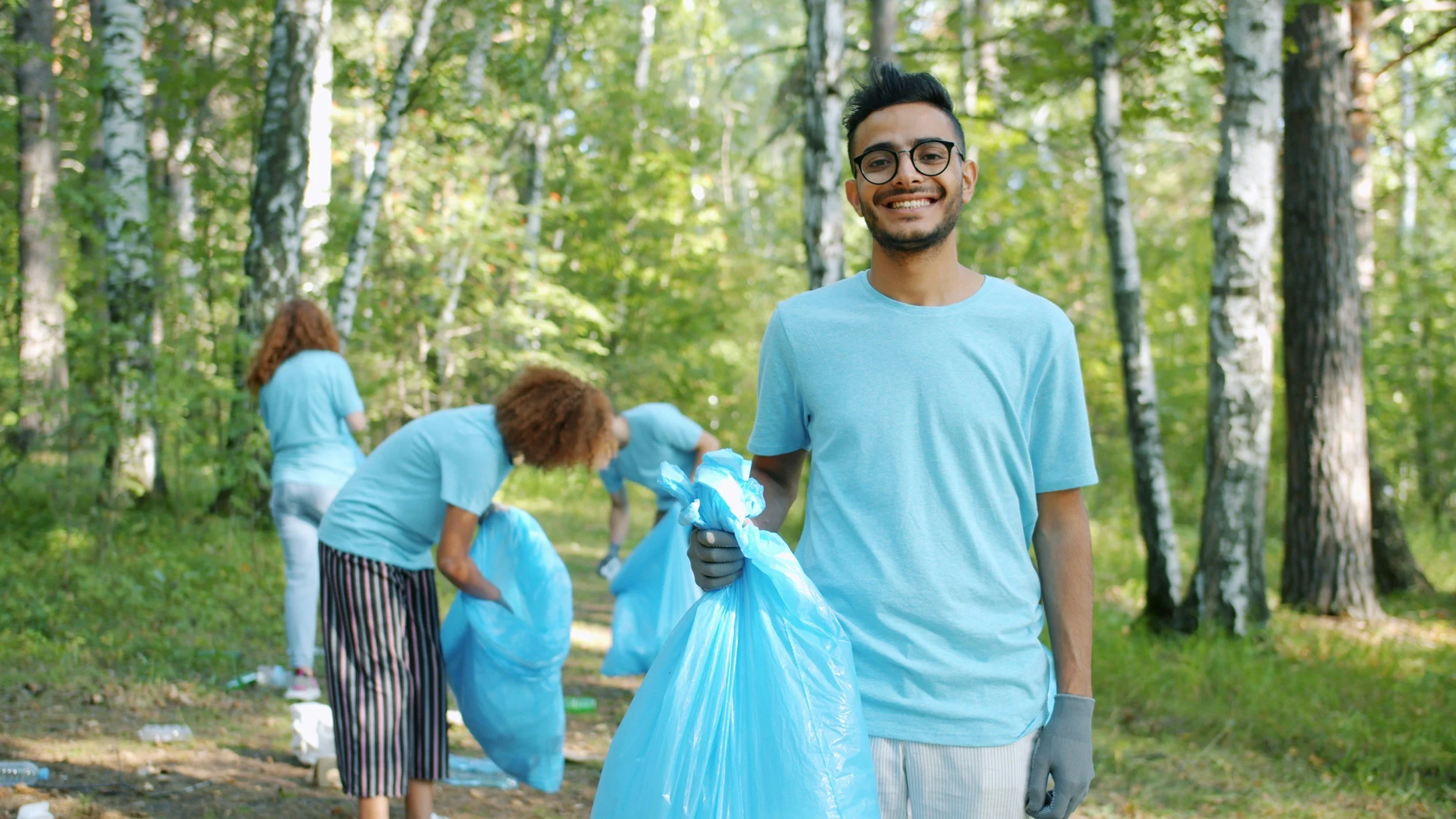 Young man with glasses smiling and holding a trash bag in a forested park while others in the background pick up trash nearby. Community Engagement, and environmental protection.