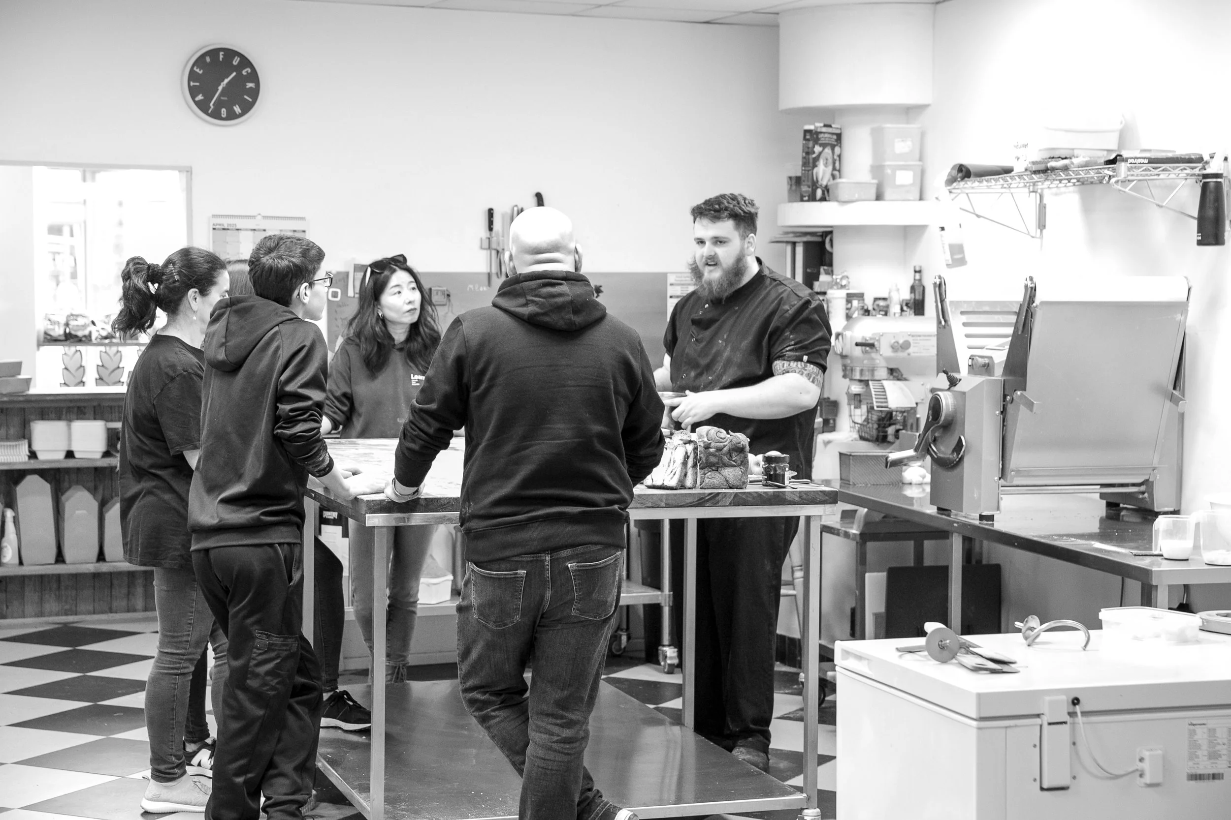 Group of five people in a commercial kitchen, three women and two men, standing around a counter with food and kitchen equipment.