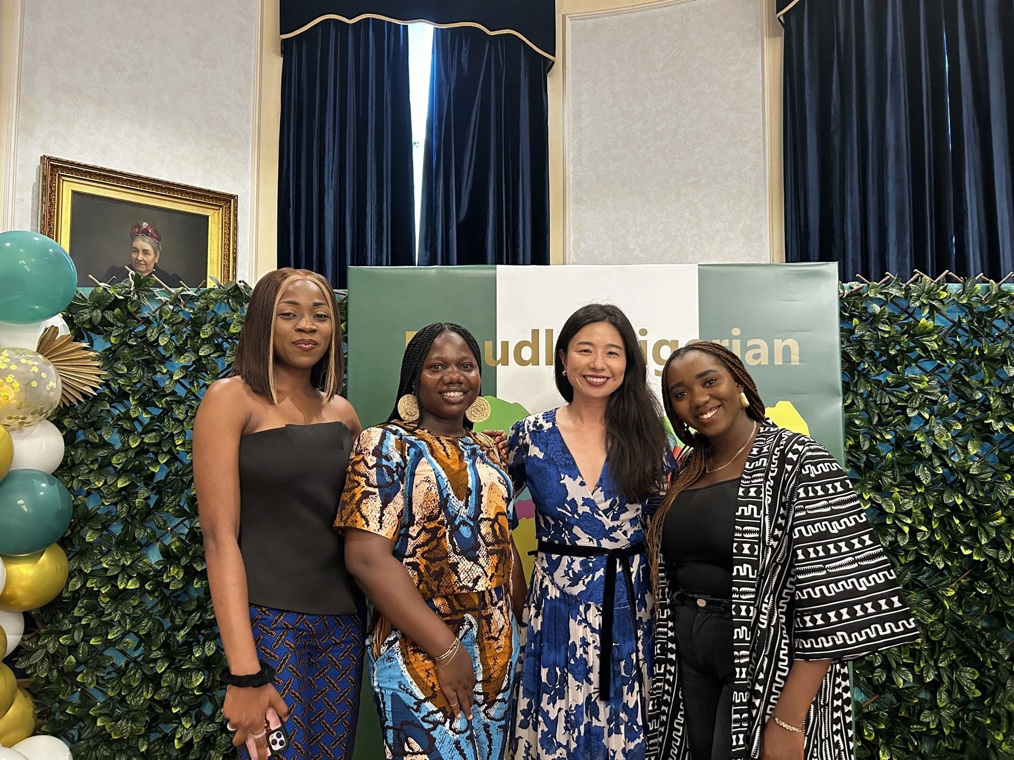 Group of five women standing together in front of a decorative backdrop with green foliage, balloons, and a portrait painting, at an indoor event.