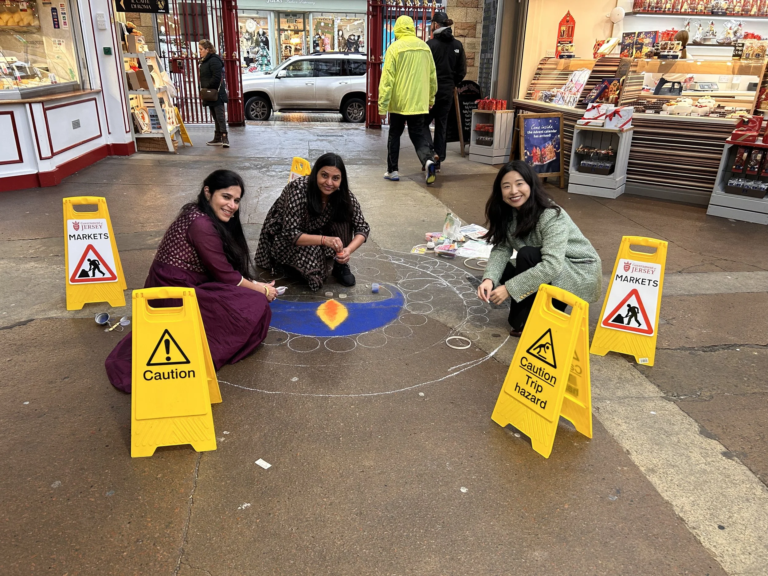 Three women sitting on the street at an outdoor market, surrounded by yellow caution signs indicating trip hazard. The women are smiling and appear to be drawing or writing on the pavement with chalk. Behind them is a shop with Christmas decorations inside and people walking by, some wearing rain jackets, and a silver car passing on the street.