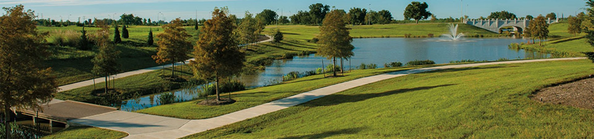 A scenic park with a lake, walking paths, green grass, trees, and a fountain in the water, with a bridge in the background.